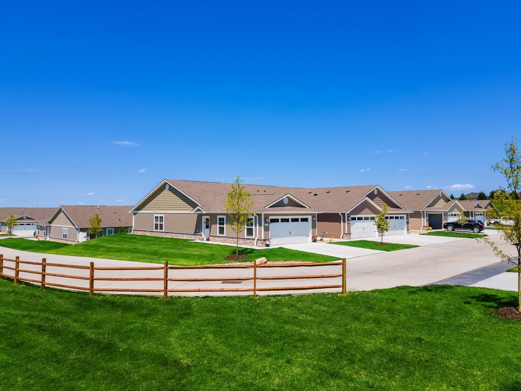 A row of single-story suburban houses with attached garages, driveways, green lawns, and a wooden fence under a clear blue sky.