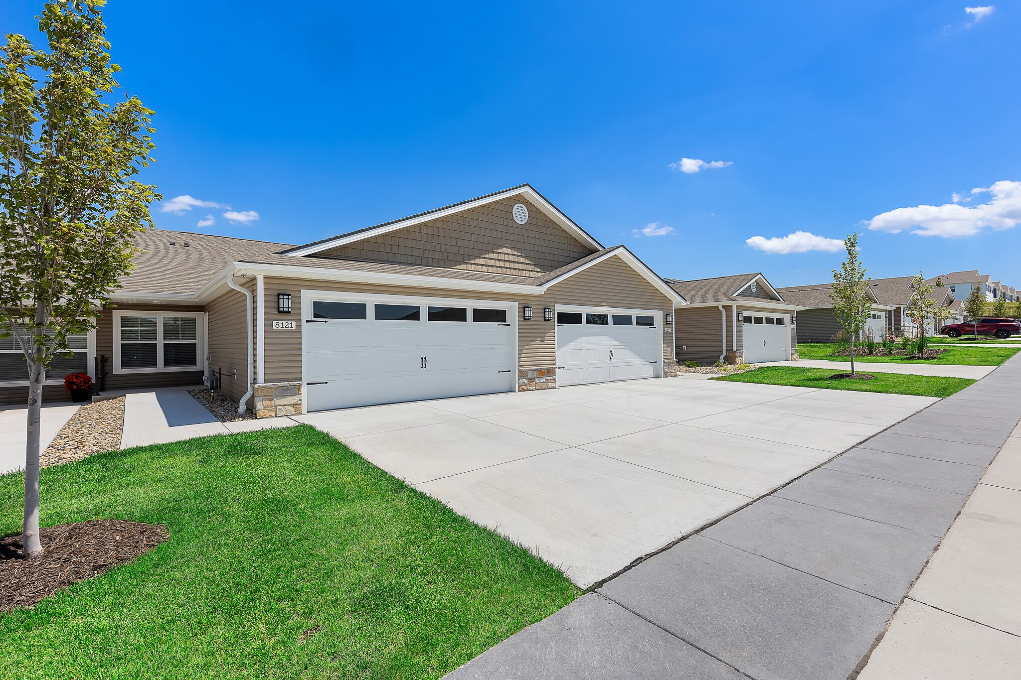 Row of single-story suburban houses with beige siding, each with double garages, concrete driveways, and small green lawns under a clear blue sky.