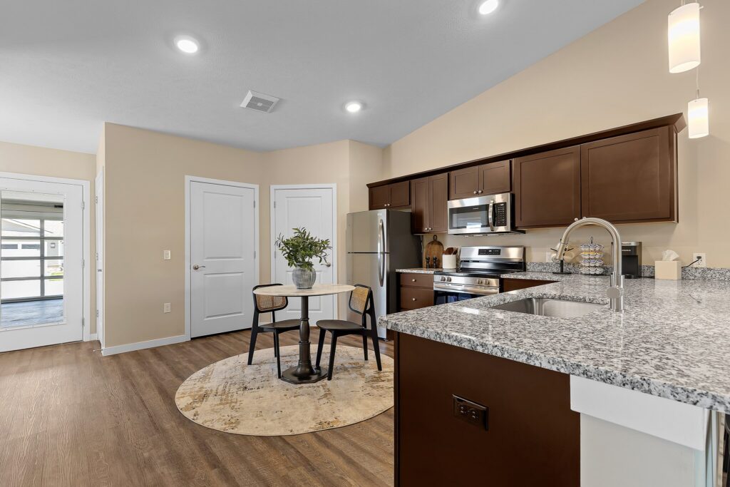 Modern kitchen with dark cabinets, stainless steel appliances, granite countertops, and a small dining table on a round rug. The space features wood flooring and neutral walls.