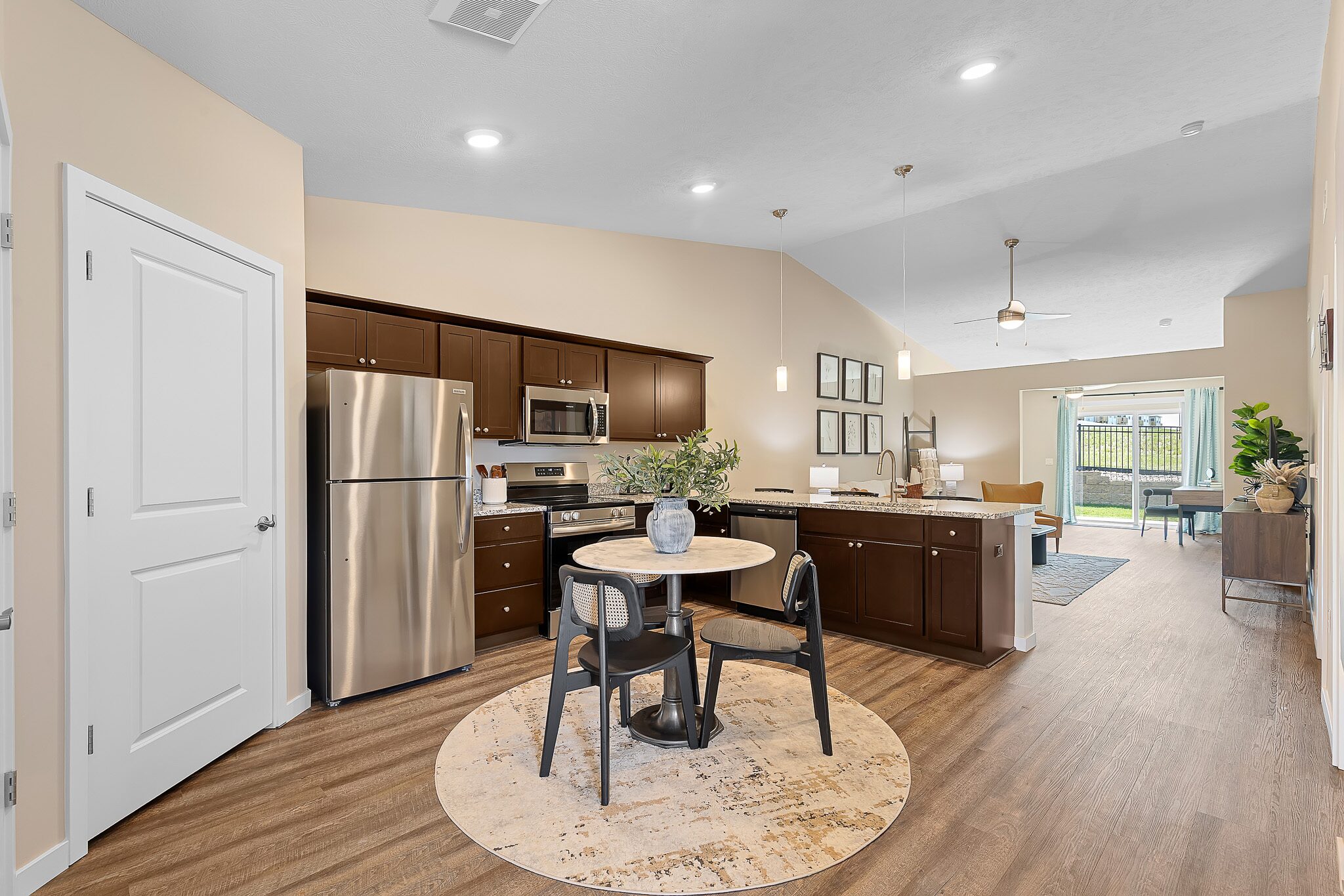 Modern kitchen with dark cabinets, stainless steel appliances, round dining table, and open layout leading to a living area with large windows and wood flooring throughout.