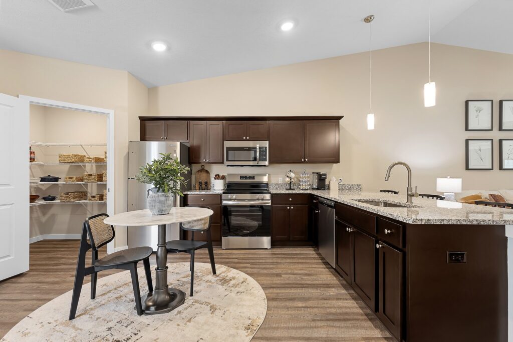 Modern kitchen with dark wood cabinets, stainless steel appliances, round dining table, and open pantry with shelves. Pendant lights hang above a granite countertop and sink.