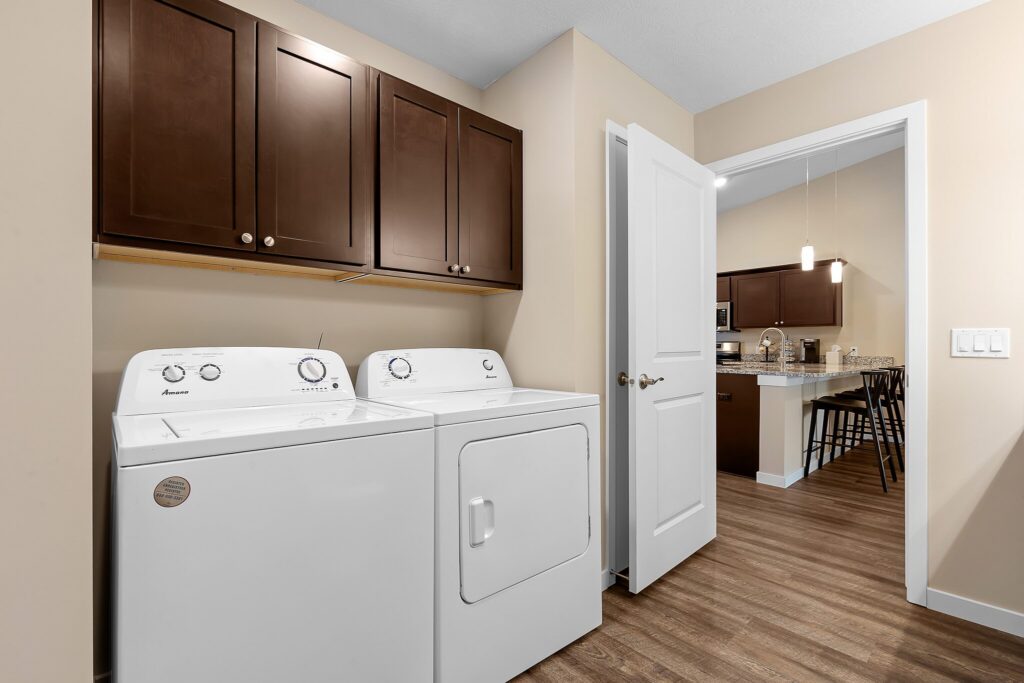 Laundry room with a white washer and dryer, brown wall cabinets above, and an open door leading to a kitchen with a bar and stools.