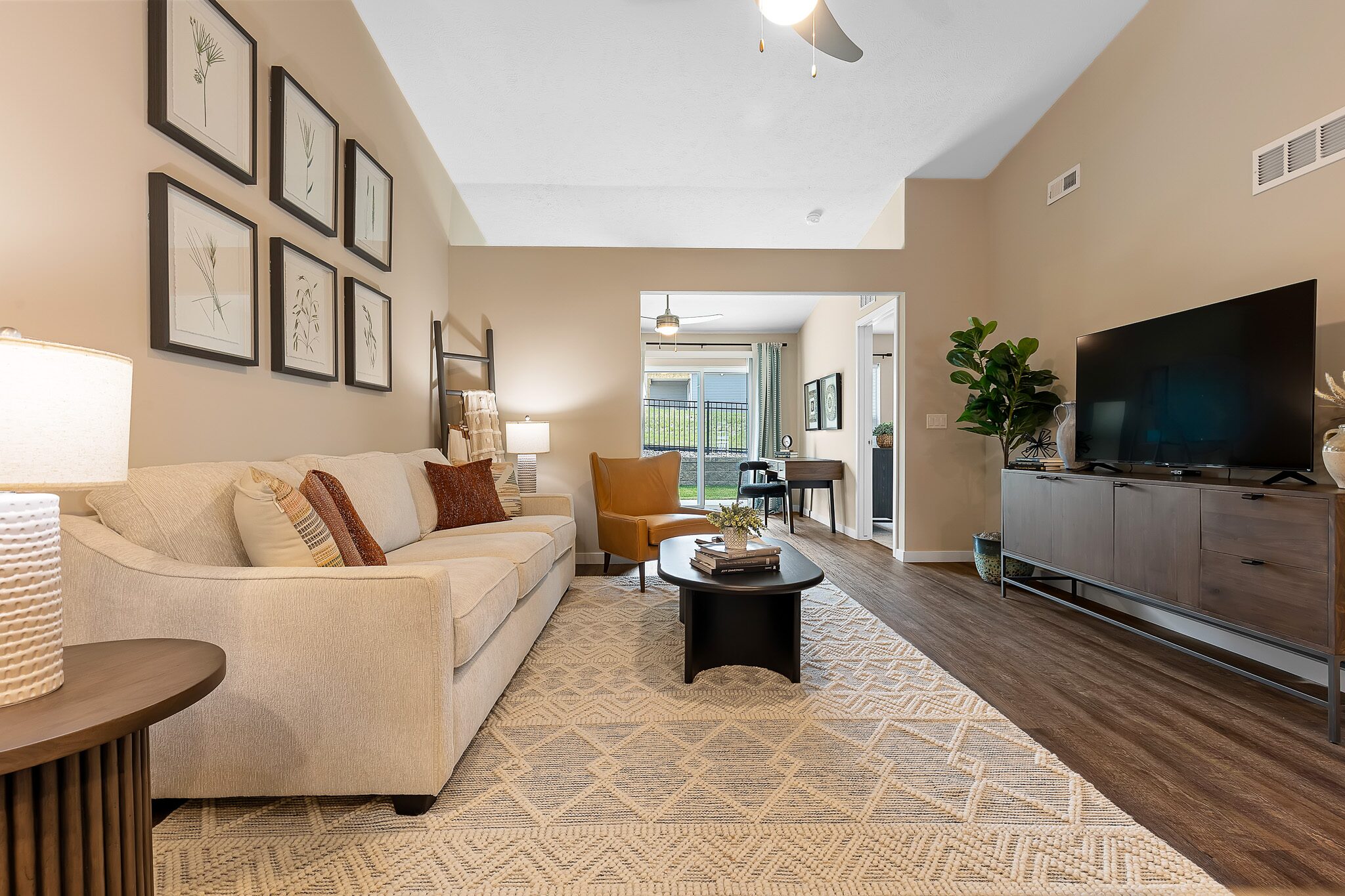 Modern living room with beige sofa, yellow chair, patterned rug, TV on console, coffee table, wall art, and view into dining area with large window and green plant.