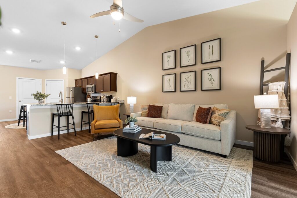 Modern living room with beige sofa, black coffee table, kitchen in background, wooden floors, neutral decor, and framed botanical prints on wall.