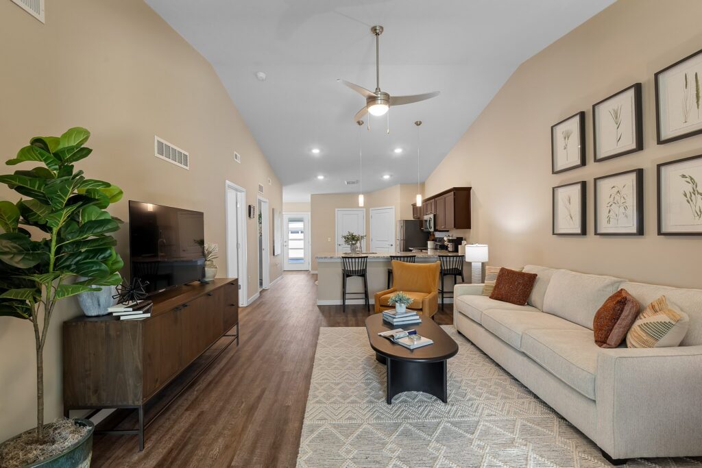 Modern living room with beige walls, a cream sofa, a TV console, wall art, and an open kitchen in the background. A potted plant and coffee table are in the foreground.