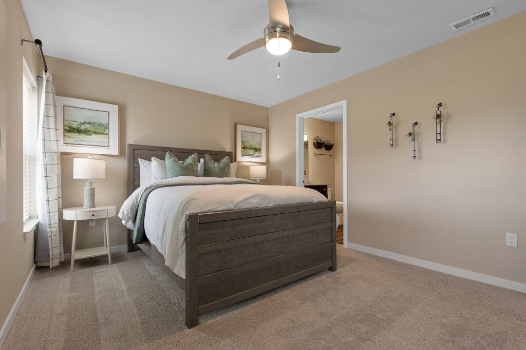 A neatly made bed with gray wooden frame, two nightstands with lamps, beige walls, carpeted floor, and a ceiling fan in a tidy bedroom.