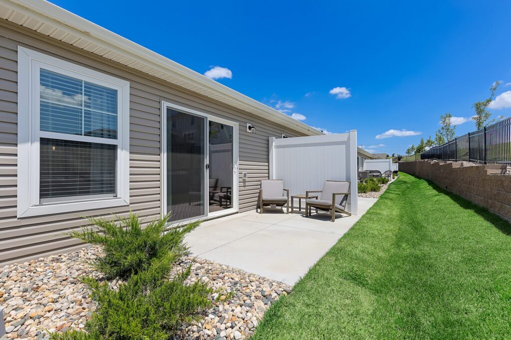 Small concrete patio with two chairs and a table, next to a beige house, sliding glass door, and green lawn under a clear blue sky.