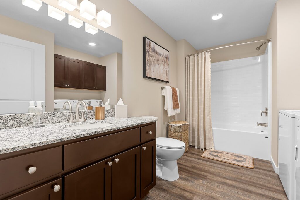 Modern bathroom with double sink vanity, granite countertop, mirror, toilet, bathtub with shower curtain, and stacked washer dryer. Wood flooring and neutral decor throughout.
