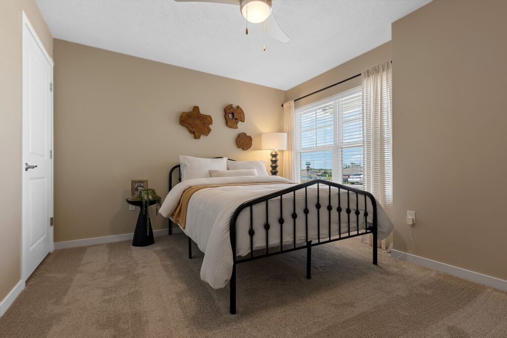 A neatly made bed with a black metal frame is centered in a beige bedroom with carpet, a lamp by the window, and wall art above the headboard.