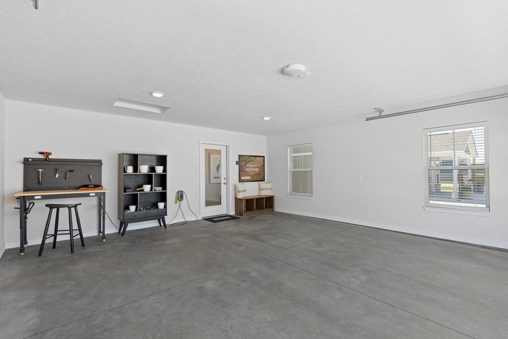 Spacious garage with gray concrete floor, workbench, shelving unit, and two windows. A door leads to the interior, and a small bench with a cushion is against the far wall.