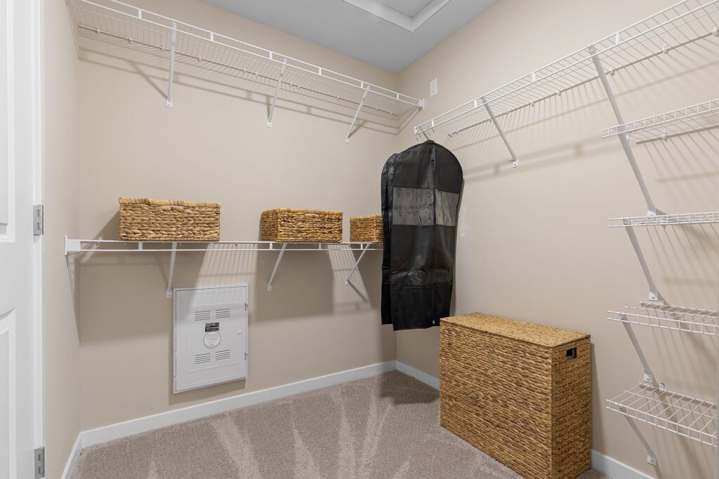 Walk-in closet with white wire shelves, three woven baskets, a black garment bag hanging, and a woven hamper on the carpeted floor. The walls are beige and the room is well-lit.