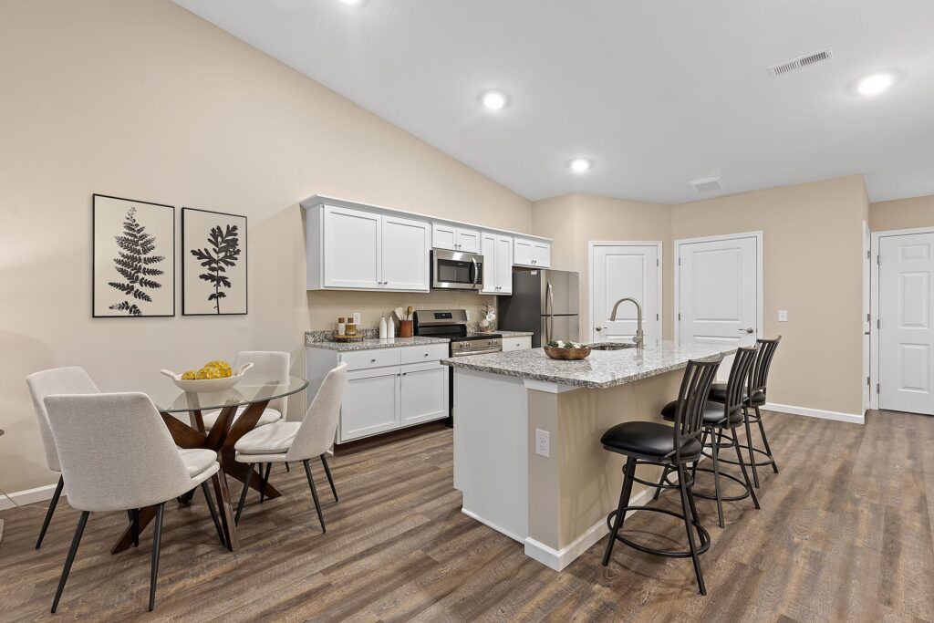 Modern kitchen with white cabinets, stainless steel appliances, a granite island with bar stools, and a small round dining table with four chairs on wood flooring.