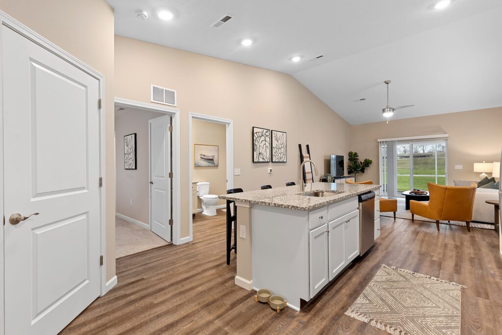 Open-concept living space with kitchen island, granite countertops, living area with sofa and TV, visible bathroom, and large window overlooking a grassy yard.