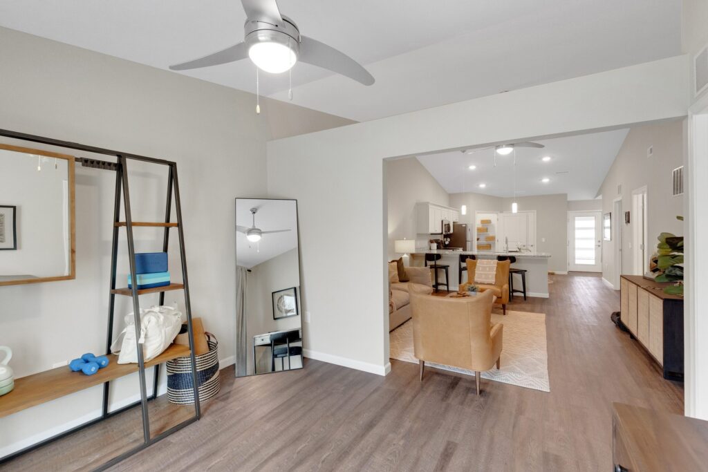 Modern open-plan living area with wood flooring, shelving, a leaning mirror, and neutral-toned furniture. Kitchen and dining area are visible in the background. Ceiling fans are installed.