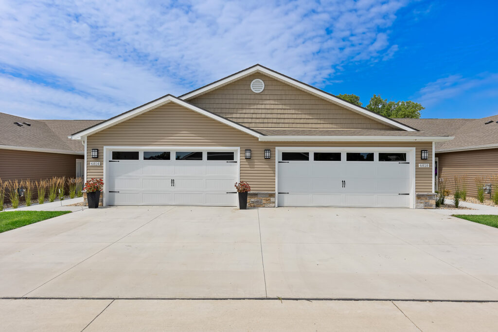 Two attached garage doors on a tan duplex with white trim, stone accents, potted plants, and a wide concrete driveway under a blue sky with scattered clouds.