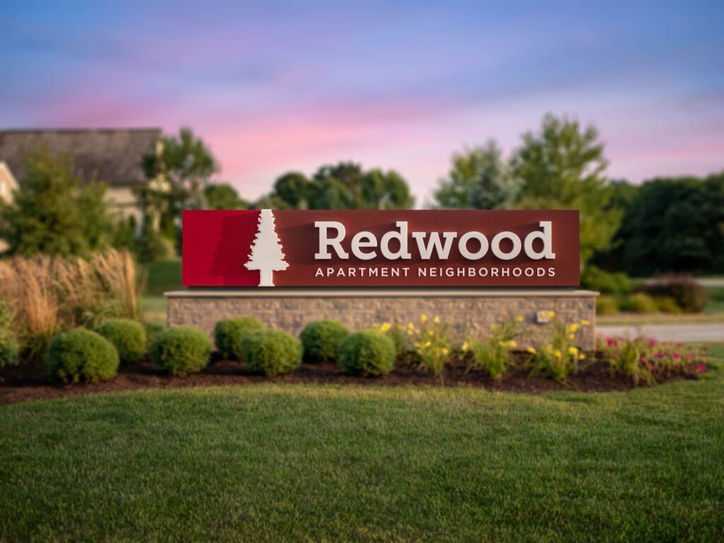 A stone sign with “Redwood Apartment Neighborhoods” and a tree logo stands on a landscaped area with grass, bushes, and flowers.