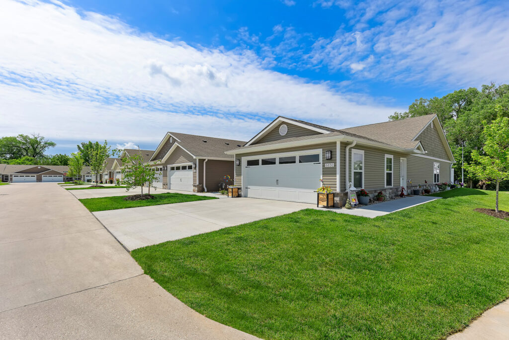 Row of single-story suburban houses with attached garages, concrete driveways, and well-kept lawns under a partly cloudy sky.
