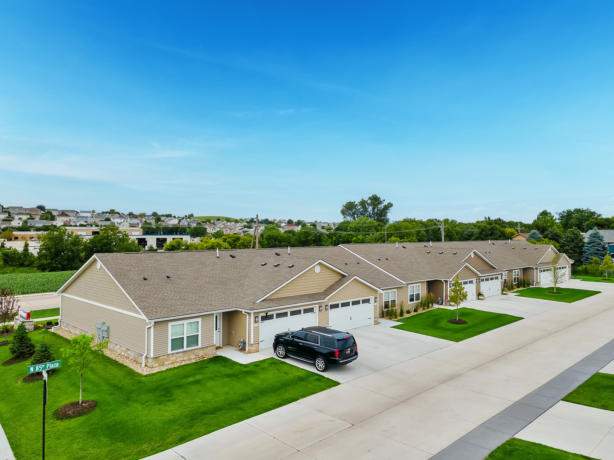 Row of single-story tan duplexes with attached garages, driveways, and green lawns on a clear day; one black SUV parked outside.