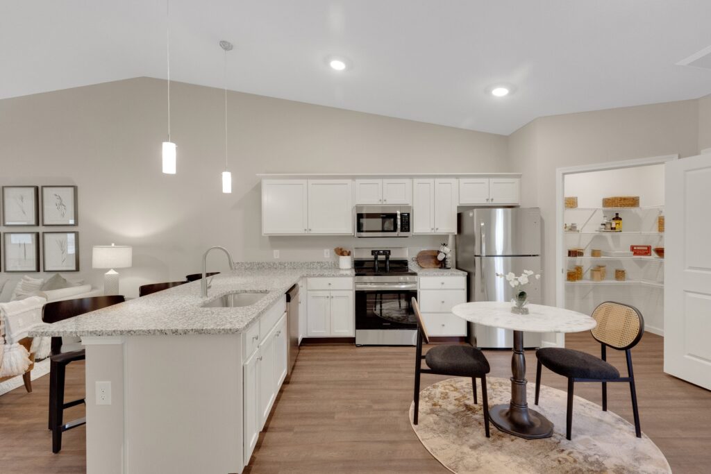 Modern kitchen with white cabinets, stainless steel appliances, a round dining table with two chairs, open shelving with baskets, and pendant lights over a granite island.