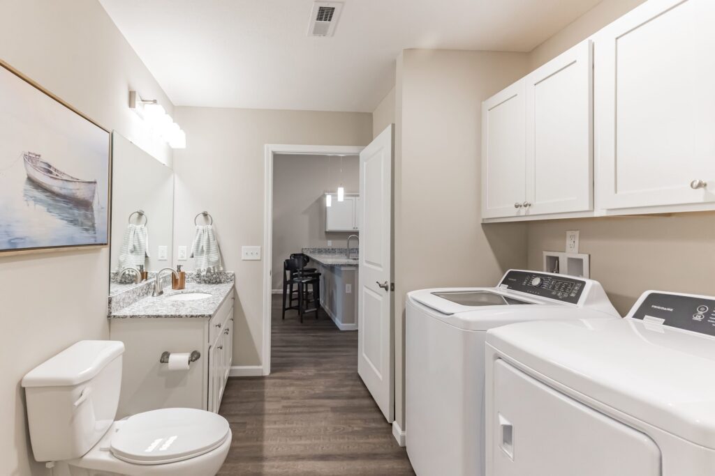 Bathroom with double vanity, toilet, washing machine, and dryer. White cabinets above appliances, wood flooring, and a boat painting on the wall. Open door shows a glimpse of another room.