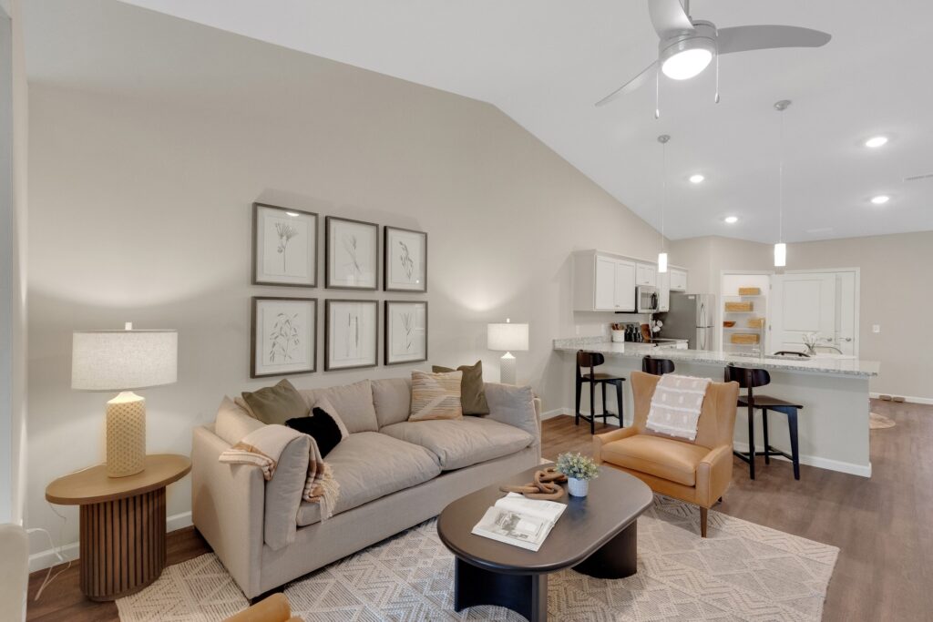 Modern living room with beige walls, sofa, and chairs, featuring framed botanical art, coffee table with decor, and open kitchen with bar stools in the background. Ceiling fan and pendant lights above.