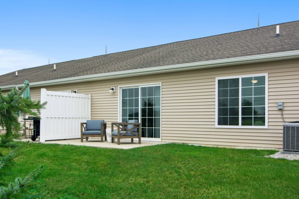 A small backyard patio with two chairs, a table, and a white fence, next to a beige house with sliding glass doors and windows.