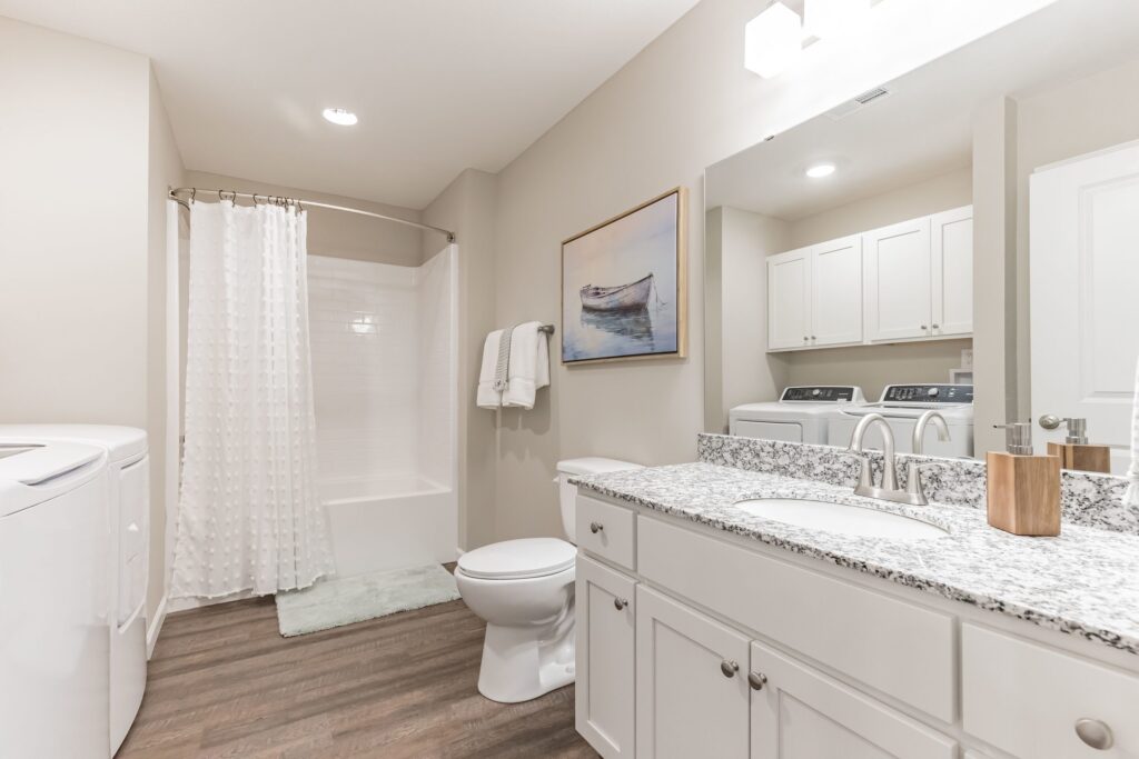 Bathroom with white cabinets, a granite countertop, a toilet, a bathtub with a shower curtain, washer and dryer, towel racks, and a framed boat picture on the wall.