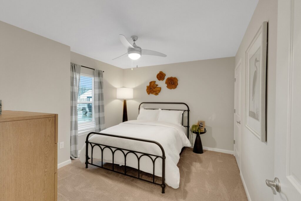 A bedroom with a metal bed frame, white bedding, a wooden dresser, window with curtains, lamp, and wall art above the headboard.
