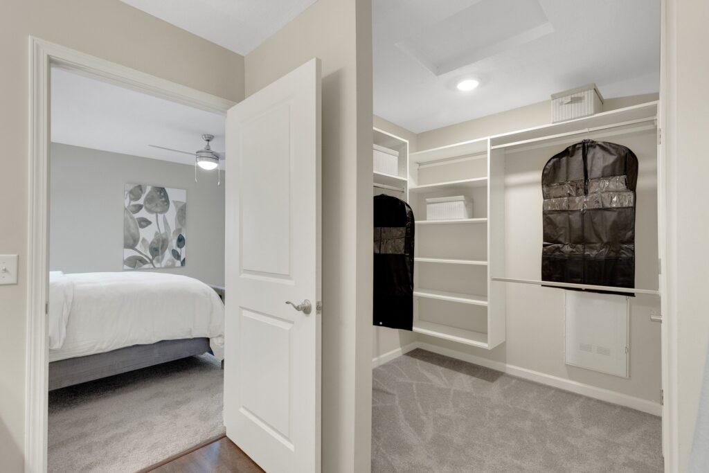 Open walk-in closet with shelves and hanging storage, viewed from an open door, adjacent to a bedroom with a made bed and wall art visible.