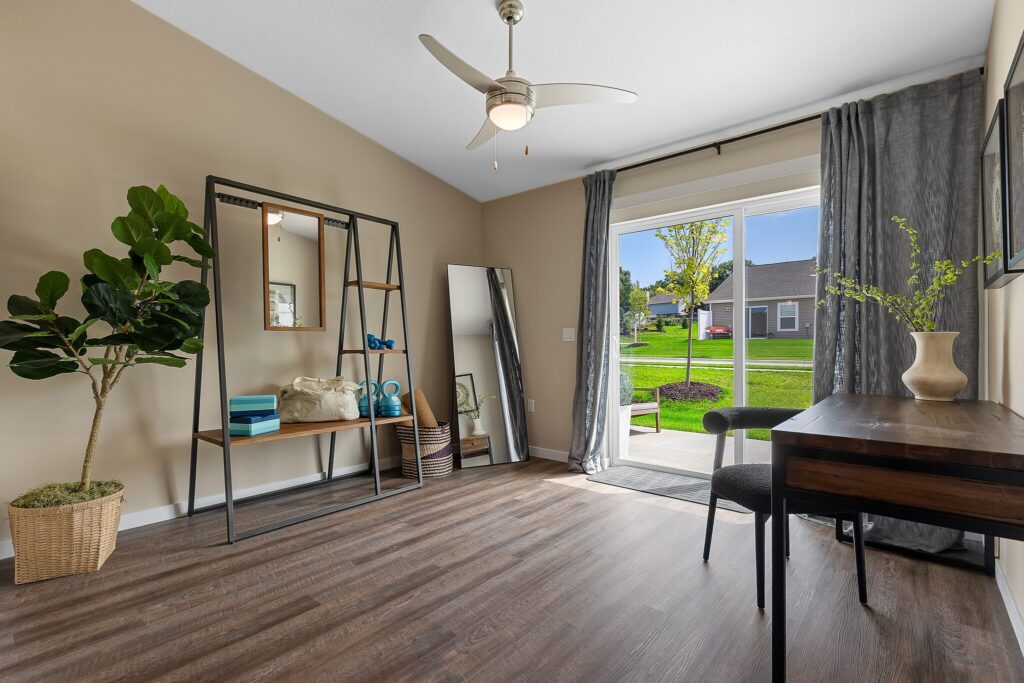 Modern home office with a desk, chair, large window with curtains, indoor plant, and shelving unit. A full-length mirror stands by the wall. Wood flooring and neutral walls.