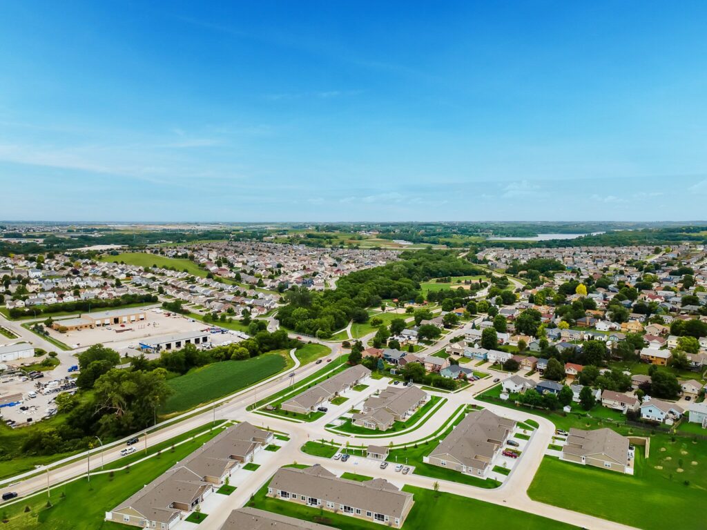 Aerial view of a suburban neighborhood with houses, roads, trees, and commercial buildings under a clear blue sky.