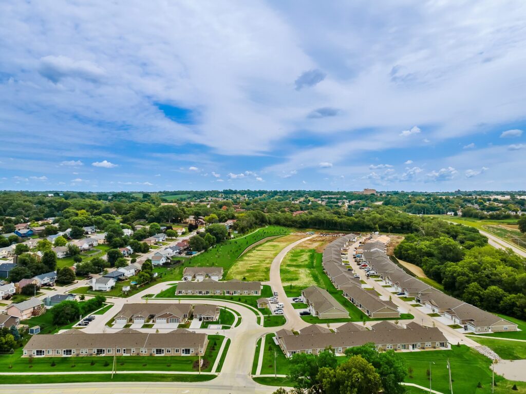 Aerial view of a suburban neighborhood with rows of houses, green lawns, curved streets, and trees under a partly cloudy sky.
