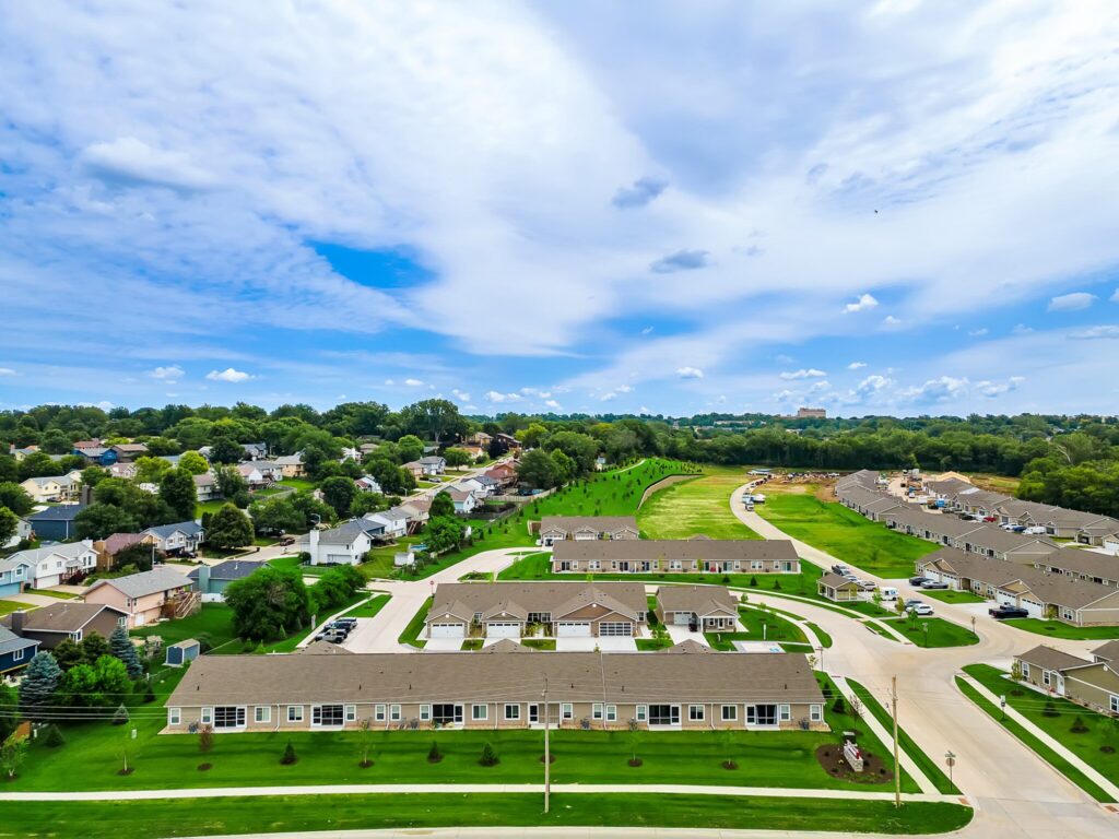 Aerial view of a suburban neighborhood with rows of houses, green lawns, trees, and a partly cloudy sky.