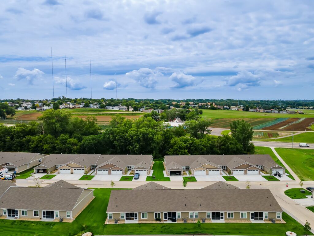 Aerial view of a suburban neighborhood with rows of houses, green lawns, trees, and farmland in the background under a partly cloudy sky.