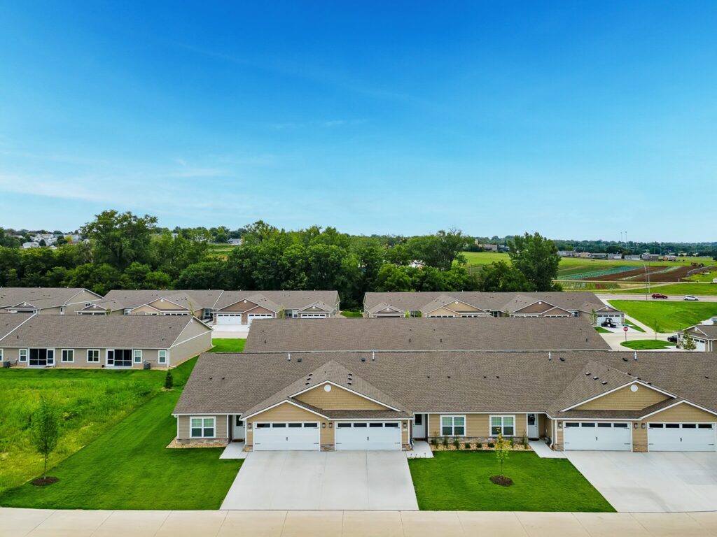 Aerial view of suburban single-story townhouses with garages, neatly mowed lawns, and a background of trees and open land under a clear blue sky.
