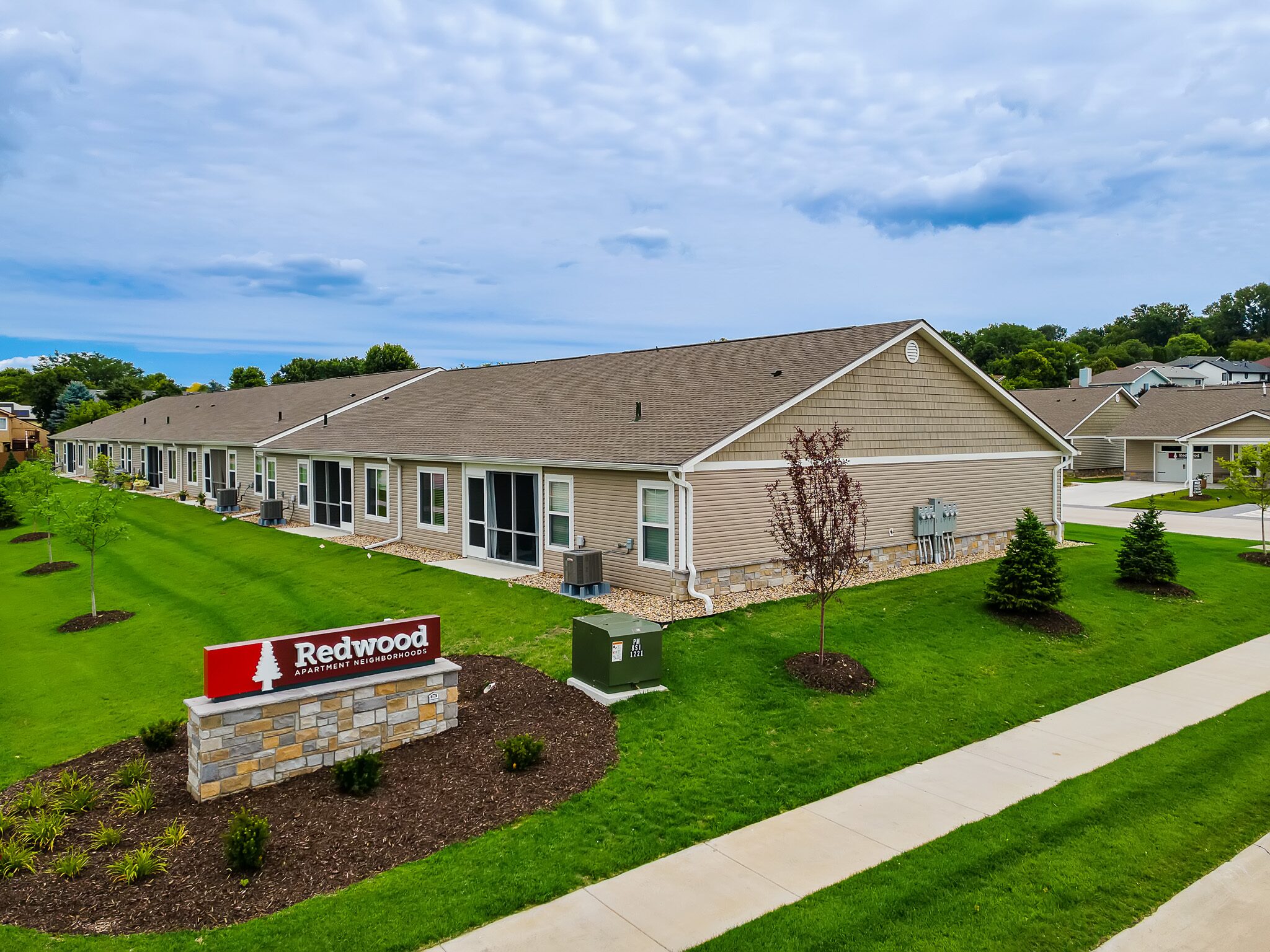 Single-story beige apartment buildings with gabled roofs, manicured lawns, and a red “Redwood” sign in front, under a partly cloudy sky.