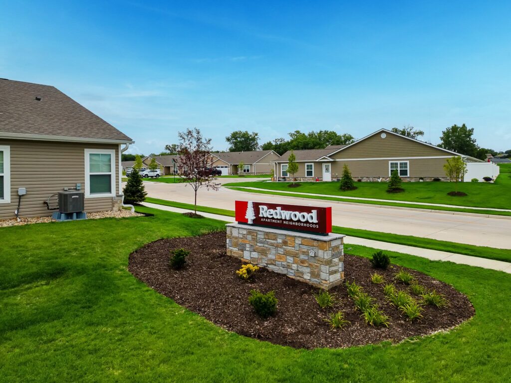 A landscaped yard with a “Redwood Apartment Neighborhoods” sign in front of single-story residential buildings under a clear blue sky.