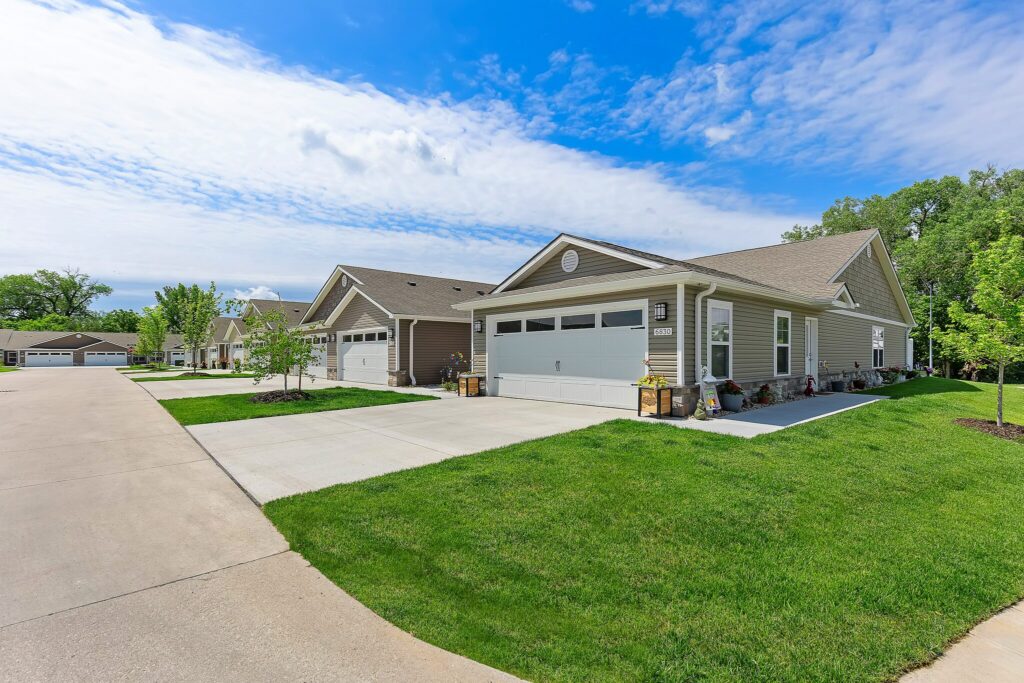 Row of modern single-story houses with attached garages, manicured lawns, and a wide paved driveway under a partly cloudy sky.