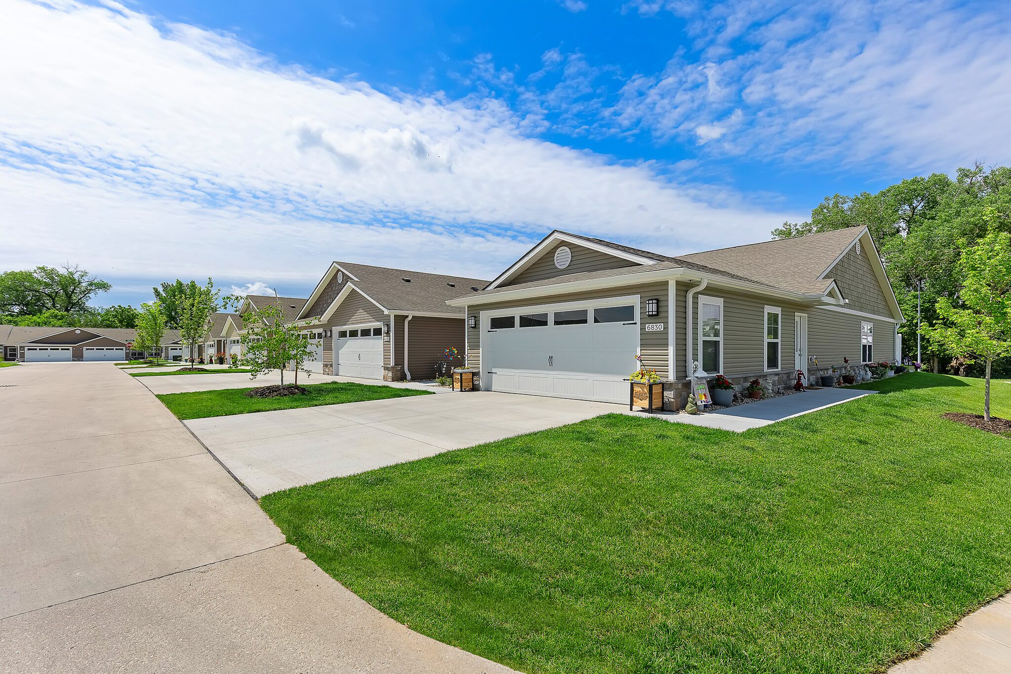 Row of modern single-story houses with attached garages, manicured lawns, and a wide paved driveway under a partly cloudy sky.