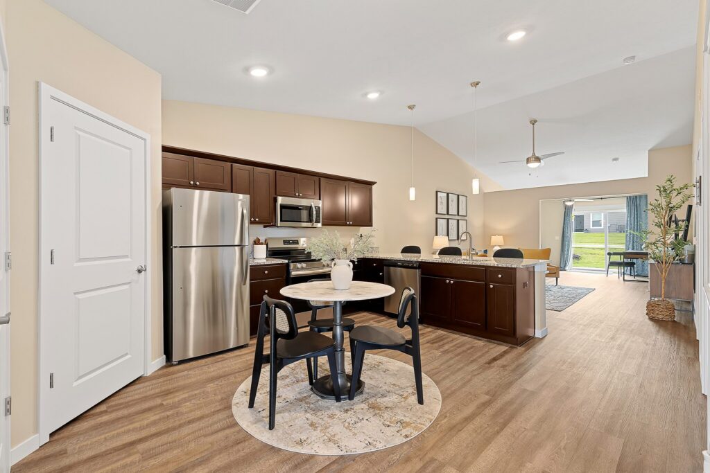 Open kitchen and dining area with dark wood cabinets, stainless steel appliances, round table with chairs, light walls, and wood flooring leading to a living area with large windows.