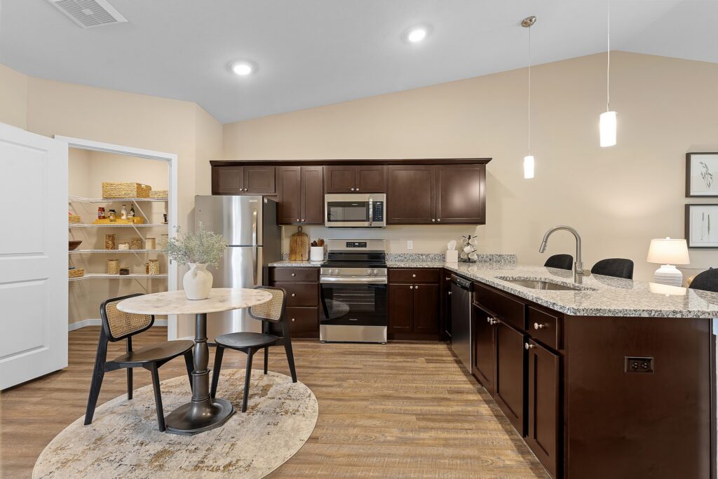 Modern kitchen with dark wood cabinets, stainless steel appliances, granite countertops, pendant lighting, and a small dining table with two chairs on a rug. Open pantry door on the left.