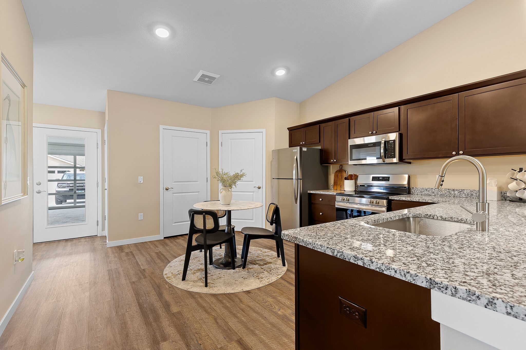 Modern kitchen with granite countertops, stainless steel appliances, dark wood cabinets, and a small dining table with four chairs on a round rug. Light wood flooring and beige walls.