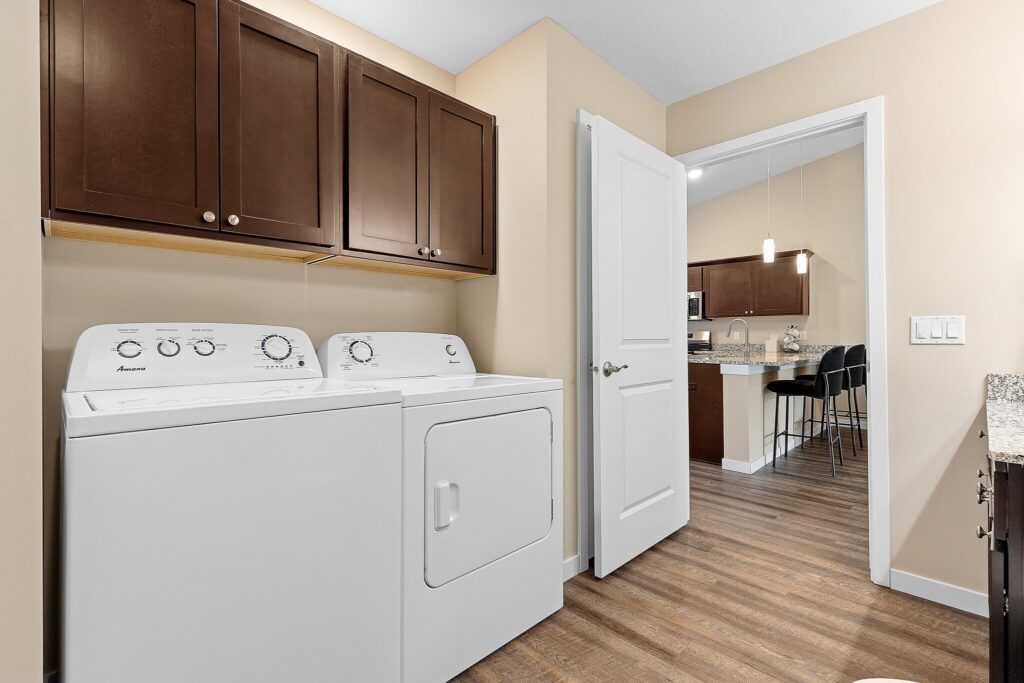 Laundry room with a white washer and dryer, brown cabinets above, open door leading to a kitchen with barstools, and wood-style flooring throughout.