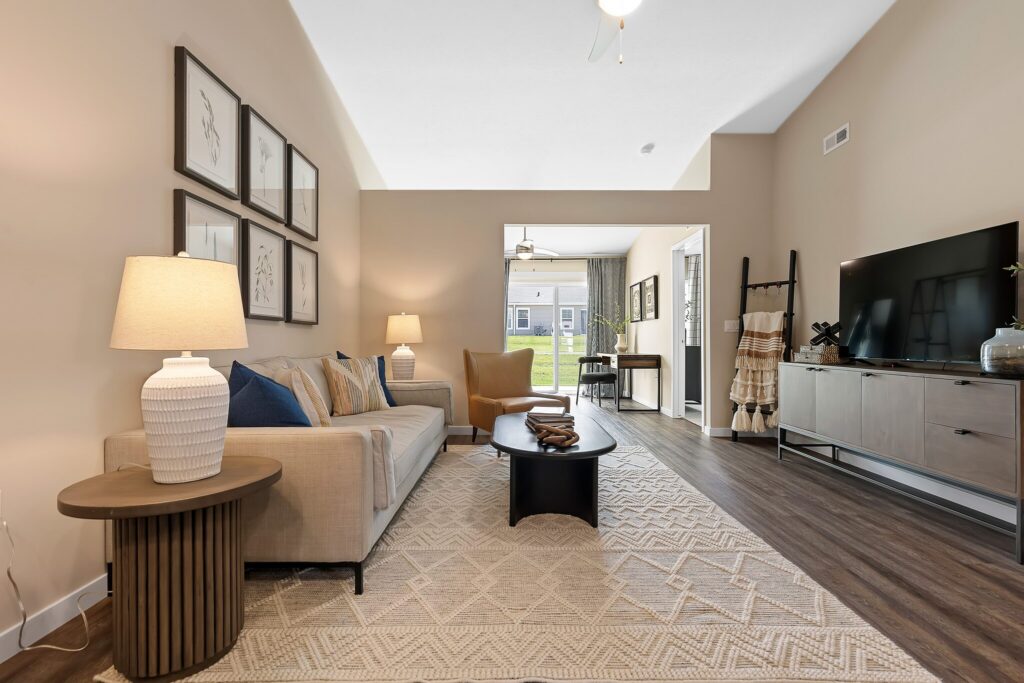 Modern living room with beige walls, neutral furniture, a patterned rug, wall art, lamps, a TV on a console, and large window providing natural light and view of a backyard.