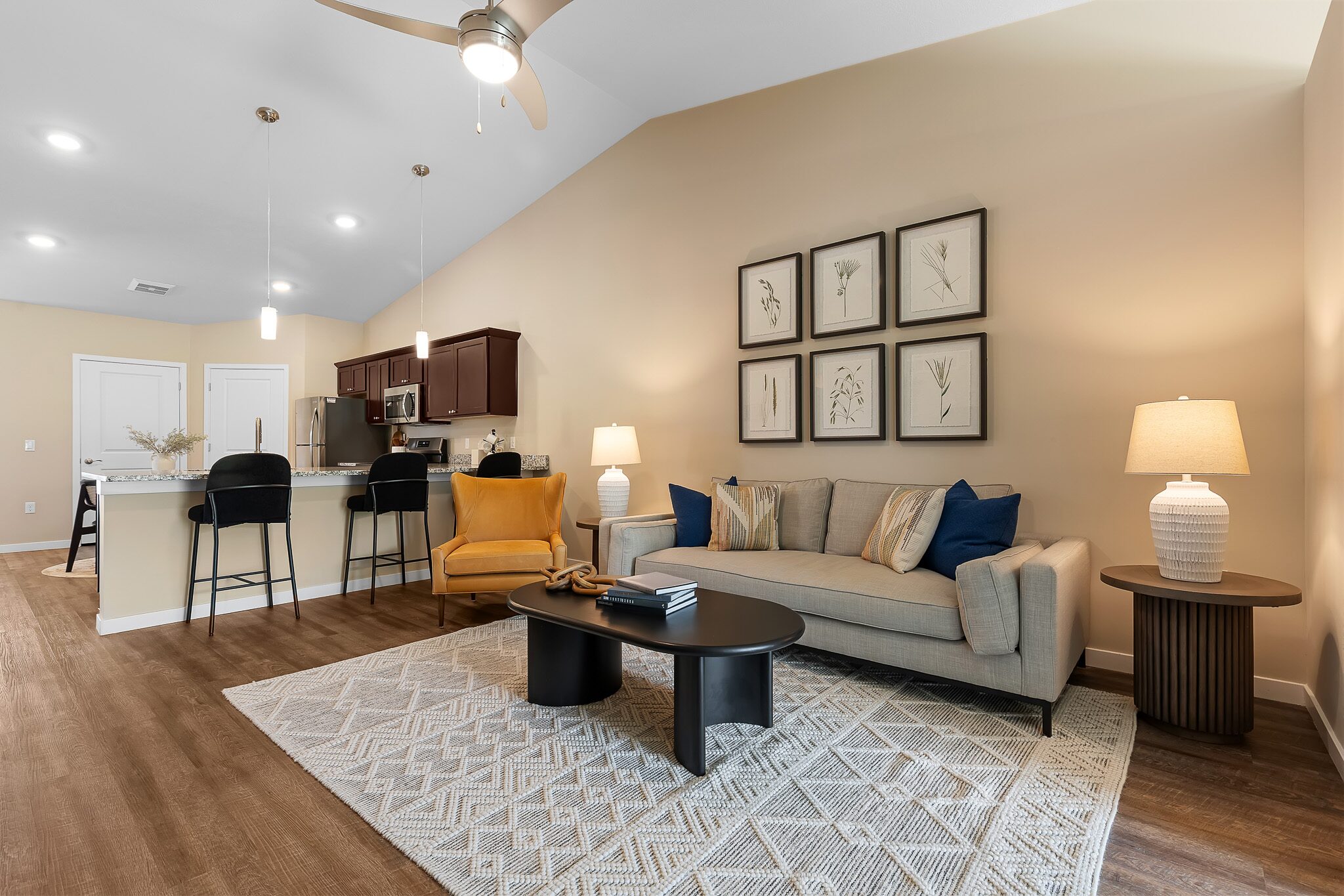 Open living room with beige walls, grey sofa, yellow armchair, black coffee table, kitchen island with bar stools, wood flooring, and framed art above the couch.