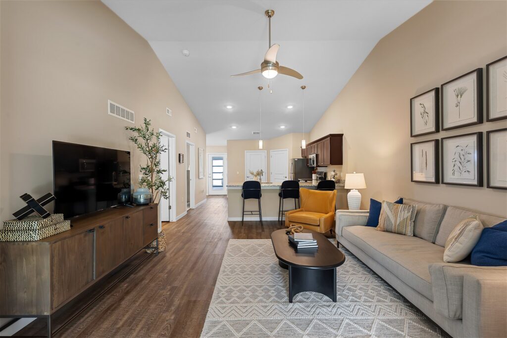 Modern living room with a beige sofa, yellow armchair, wall art, TV console, and open kitchen; wood floors and neutral walls create a warm, inviting atmosphere.