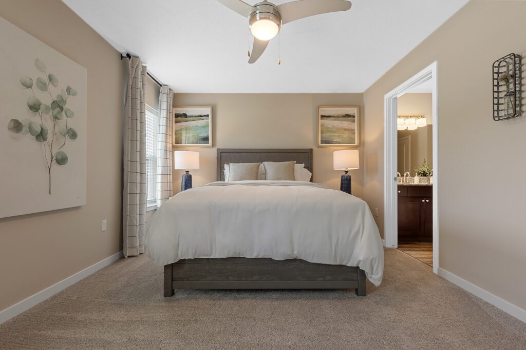 A modern bedroom with a neatly made bed, two bedside tables with lamps, neutral walls, a ceiling fan, and an adjacent bathroom visible through an open door.