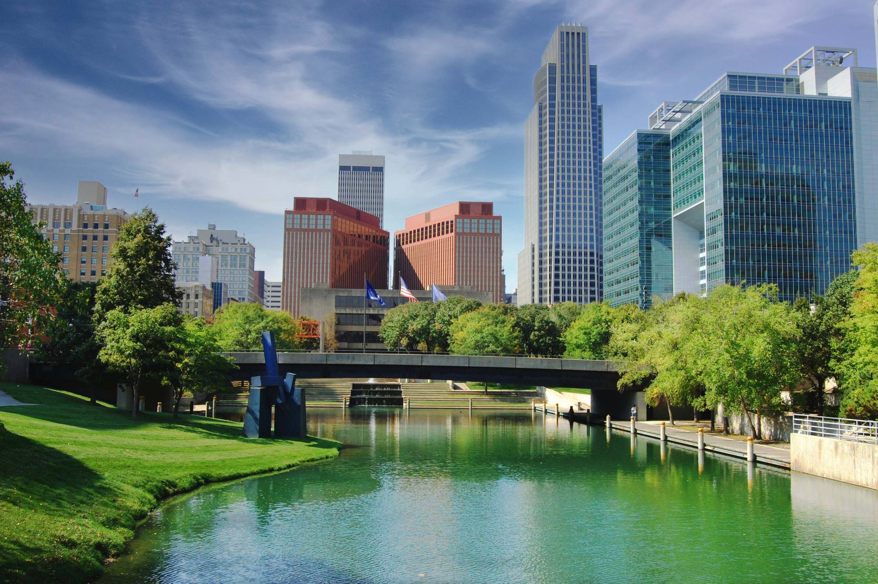 A city skyline with modern buildings, a green park, and a small river running under a bridge on a sunny day.