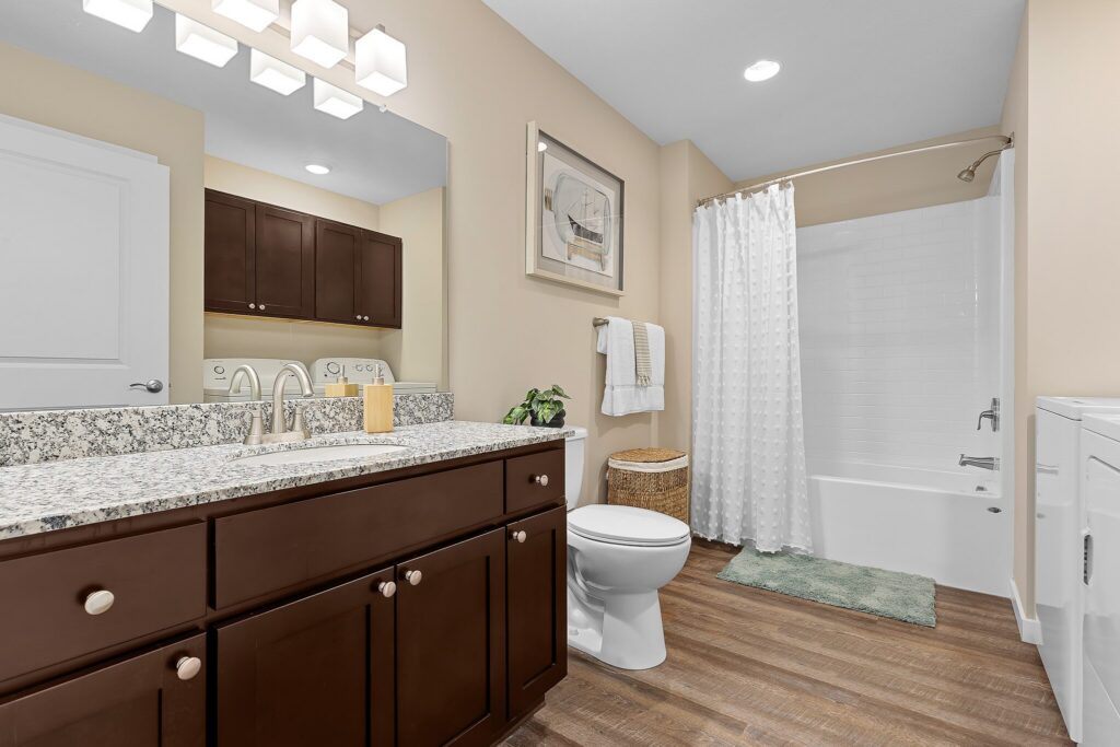 Modern bathroom with dark wood vanity, granite countertop, toilet, white bathtub with shower, and light wood flooring. White walls and decor, with towels and a green bath mat.