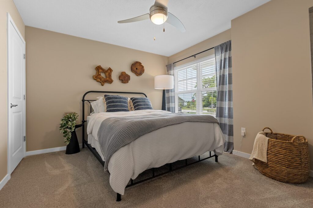 A neatly made bed with striped pillows, a ceiling fan, a lamp by the window, wall decor, a plant in a black vase, and a wicker basket on a beige carpeted floor.